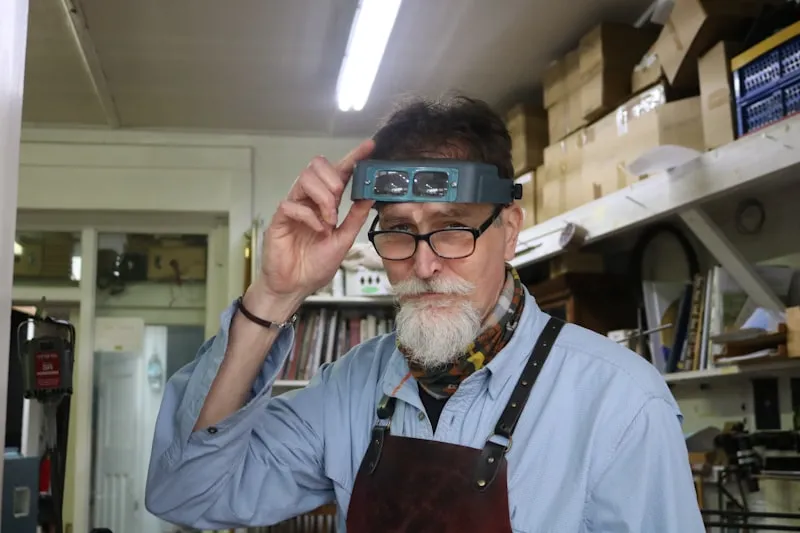 A master craftsman in a leather apron adjusting a head-mounted magnifying visor inside a custom fabrication workshop.